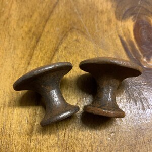 May include: Two antique, weathered metal cabinet knobs. Each knob has a rounded top and a flared base, with a dark, rusty brown finish. The knobs are displayed on a wooden surface.