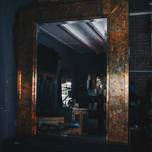 May include: A large, ornate mirror with a copper-toned frame. The frame has a textured, aged look, with hints of rust and patina. The mirror reflects a dimly lit room with various objects and furniture.