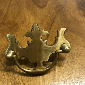 Könnte beinhalten: Ein messingfarbener Schubladengriff mit einem verzierten, symmetrischen Design. Der Griff hat eine dekorative Oberseite mit einem gebogenen Griff und zwei abgerundeten Endstücken. Die Hardware befindet sich auf einer Holzoberfläche.