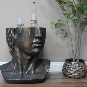 May include: A bronze-effect bust of a classical figure forms a side table. A clear glass bottle and a small glass sit on the table. A decorative plant in a textured pot is to the right.