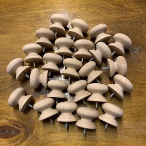 May include: A pile of unfinished wooden knobs with screws attached. The knobs have a rounded top and a flared base, and are arranged on a wooden surface. The natural wood colour and simple design suggest a craft or DIY project.