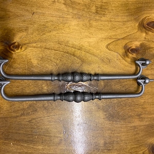 May include: Two dark grey metal drawer pulls with a textured centre and curved handles. The pulls are set against a wooden surface, highlighting their design and potential use in furniture restoration or new builds.