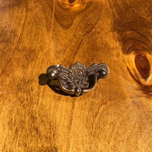 May include: A brass cabinet pull with a floral design. The pull has a curved shape and two small knobs on the sides.