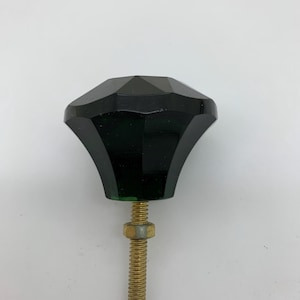May include: A dark green glass cabinet knob with a faceted design and a brass screw. The knob has a flared base. The screw is threaded and has a brass nut. The background is plain white.