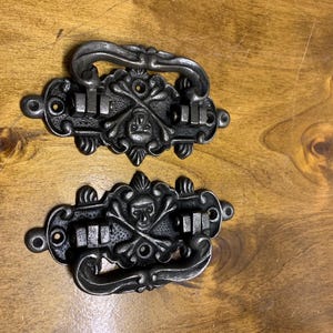 May include: Two black metal drawer pulls with a skull and crossbones design. The pulls have a decorative, ornate design.