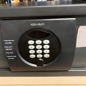 May include: A black hotel safe with a digital keypad. The keypad has numbered buttons from 0-9, a reset button, and a lock button. Instructions are printed on the safe's surface. The brand name ASSA ABLOY is visible.