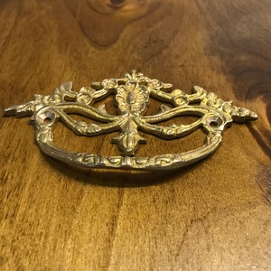 May include: A gold-toned metal drawer pull with an ornate design featuring a central floral motif and scrolling vines. The pull has two holes for mounting.