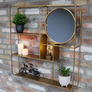 May include: A gold-coloured metal wall shelf featuring a circular mirror, shelves, and decorative items. The shelves display a small potted plant, candles, a miniature bicycle, and a small lamp. The backdrop is a brick wall.