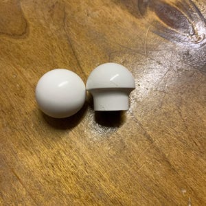May include: Two white cabinet knobs. One is a sphere, the other is a mushroom-shaped knob. Both are made of a glossy, solid white material. The knobs are set against a wooden surface.