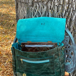 May include: A teal leather satchel bag with a brown leather lining and a zippered pocket. The bag has a flap closure and a brass buckle. The bag is open and the inside is visible.