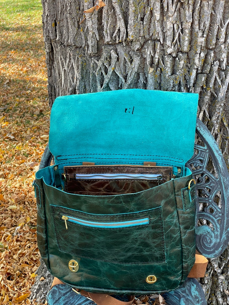 May include: A teal leather satchel bag with a brown leather lining and a zippered pocket. The bag has a flap closure and a brass buckle. The bag is open and the inside is visible.