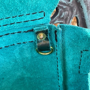 May include: Close-up of a teal leather bag with a brown leather strap and a gold metal buckle. The bag has a stitched seam and a flap.