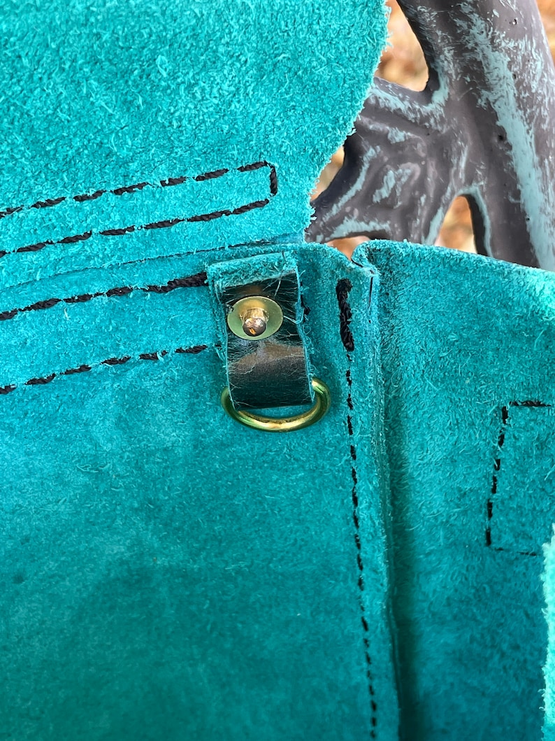 May include: Close-up of a teal leather bag with a brown leather strap and a gold metal buckle. The bag has a stitched seam and a flap.