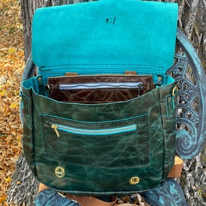 May include: A dark green leather satchel with a teal blue lining and a zippered pocket. The bag has a brown leather strap and gold metal hardware.