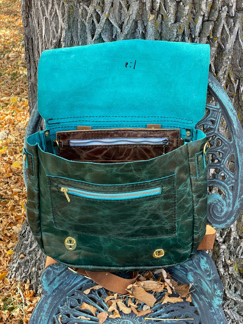 May include: A dark green leather satchel with a teal blue lining and a zippered pocket. The bag has a brown leather strap and gold metal hardware.