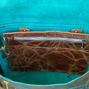 May include: The inside of a teal leather bag with a brown leather zippered pocket. The bag has a flap closure and a zippered pocket on the inside.
