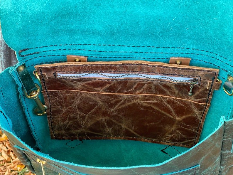 May include: The inside of a teal leather bag with a brown leather zippered pocket. The bag has a flap closure and a zippered pocket on the inside.