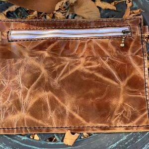 May include: A brown leather pouch with a zipper closure and a small metal charm. The pouch has a distressed look and is made of high-quality leather.