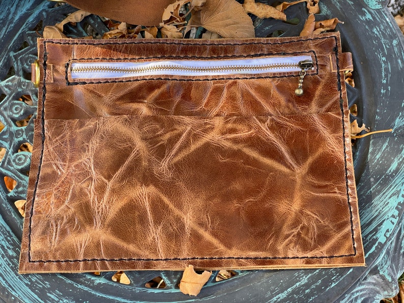 May include: A brown leather pouch with a zipper closure and a small metal charm. The pouch has a distressed look and is made of high-quality leather.