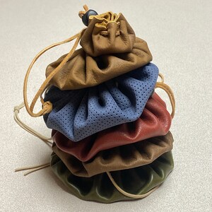 May include: A stack of five leather pouches in various colors, including brown, blue, red, and green. The pouches are all closed and have drawstrings. The top pouch has a black bead on the drawstring.
