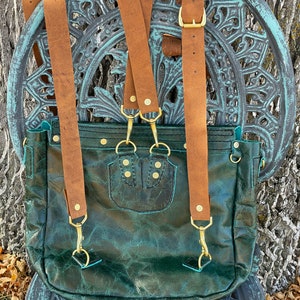 May include: A dark green leather backpack with brown leather straps and gold hardware. The backpack has a flap closure and a small pocket on the front.