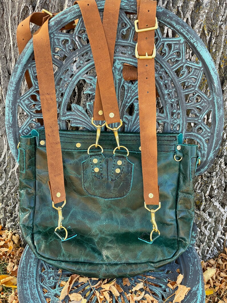 May include: A dark green leather backpack with brown leather straps and gold hardware. The backpack has a flap closure and a small pocket on the front.
