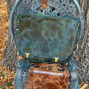 May include: A teal green leather satchel bag with a brown leather strap and a smaller brown leather pouch with a zipper. The bag is sitting on a decorative metal chair.