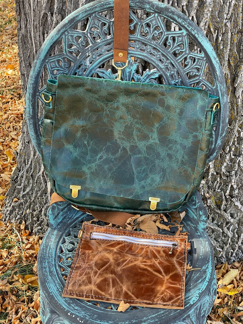 May include: A teal green leather satchel bag with a brown leather strap and a smaller brown leather pouch with a zipper. The bag is sitting on a decorative metal chair.