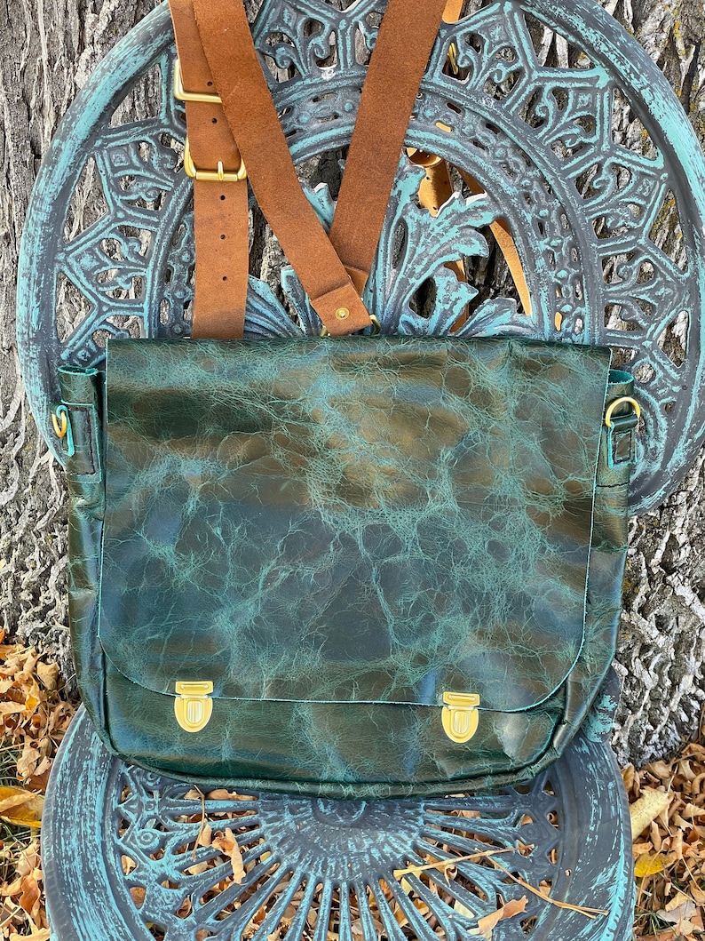 May include: A dark green leather satchel with brass buckles and brown leather straps. The bag is sitting on a decorative metal chair.