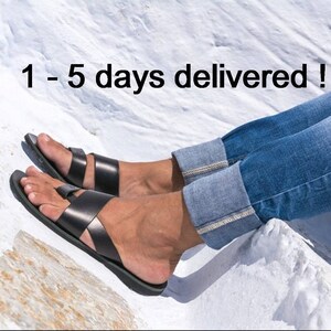 K&ouml;nnte beinhalten: Schwarze Ledersandalen mit einem Kreuzriemen-Design. Die Sandalen werden mit einer blauen Jeans getragen. Der Text "1 - 5 days delivered !" ist im Bild zu sehen.