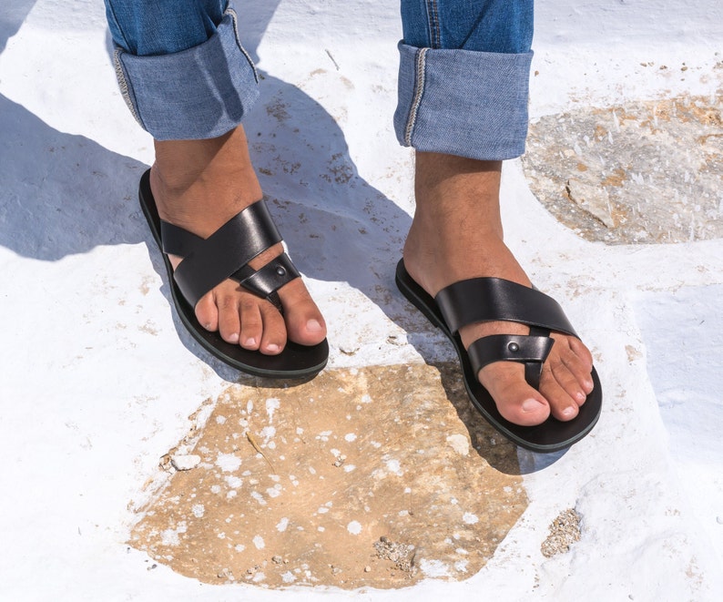 K&ouml;nnte beinhalten: Schwarze Ledersandalen mit Zehenschlaufe und Kreuzriemen-Design. Die Sandalen haben eine flache Sohle. Die Sandalen werden mit einer blauen Jeans getragen. Sie werden auf einer wei&szlig;en und beigefarbenen Steinoberfl&auml;che gezeigt.