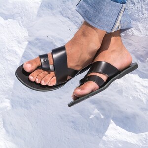 K&ouml;nnte beinhalten: Schwarze Ledersandalen mit minimalistischem Design. Die Sandalen haben eine flache Sohle und Riemen, die sich &uuml;ber dem Fu&szlig; kreuzen. Das Bild zeigt die Sandalen auf einer wei&szlig;en, strukturierten Oberfl&auml;che.
