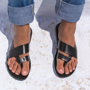 K&ouml;nnte beinhalten: Schwarze Ledersandalen mit Zehenschlaufe und gekreuzten Riemen. Die Sandalen werden mit einer blauen Jeans getragen. Das Bild ist eine Nahaufnahme der F&uuml;&szlig;e und Sandalen.