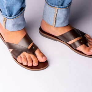K&ouml;nnte beinhalten: Dunkelbraune Ledersandalen mit einem Kreuzriemendesign. Die Sandalen haben einen Zehensteg und eine dunkelbraune Sohle. Das Bild zeigt die Sandalen an den F&uuml;&szlig;en einer Person.