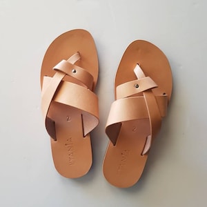 K&ouml;nnte beinhalten: Ein Paar hellbraune Ledersandalen. Die Sandalen haben einen Zehenriemen im Zehensteg-Stil und gekreuzte Riemen &uuml;ber dem Fu&szlig;. Die Sandalen sind mit dem Wort "KYANIA" und der Zahl "2" beschriftet.