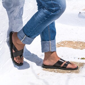K&ouml;nnte beinhalten: Schwarze Ledersandalen mit einem Kreuzriemen-Design. Die Sandalen werden mit blauen Jeans mit hochgekrempelten Manschetten getragen. Das Bild ist im Freien auf einer wei&szlig;en Oberfl&auml;che aufgenommen.