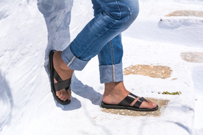 K&ouml;nnte beinhalten: Schwarze Ledersandalen mit einem Kreuzriemen-Design. Die Sandalen werden mit blauen Jeans mit hochgekrempelten Manschetten getragen. Das Bild ist im Freien auf einer wei&szlig;en Oberfl&auml;che aufgenommen.