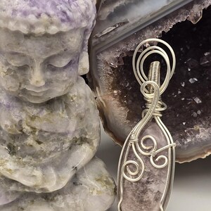 May include: A handcrafted pendant with a light purple gemstone wrapped in silver wire. The pendant is displayed next to a carved purple Buddha statue and a geode.