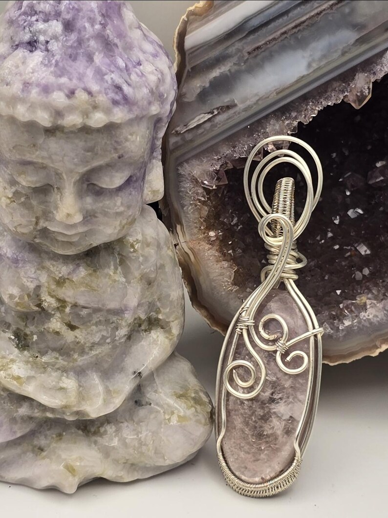 May include: A handcrafted pendant with a light purple gemstone wrapped in silver wire. The pendant is displayed next to a carved purple Buddha statue and a geode.