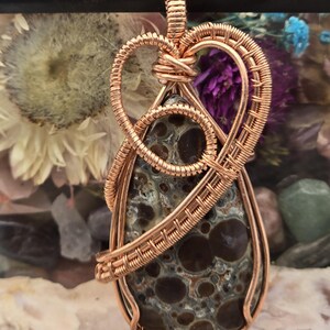 May include: A copper wire-wrapped pendant featuring a teardrop-shaped stone with a speckled pattern in shades of brown, gray, and cream. The copper wire is intricately woven around the stone, forming a decorative bail and heart-shaped design.