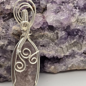 May include: A handcrafted pendant featuring a polished, oval-shaped, lavender-colored stone encased in a silver wire frame. The wirework includes decorative swirls and a wrapped top, creating a unique jewelry piece. The background is a purple crystal formation.