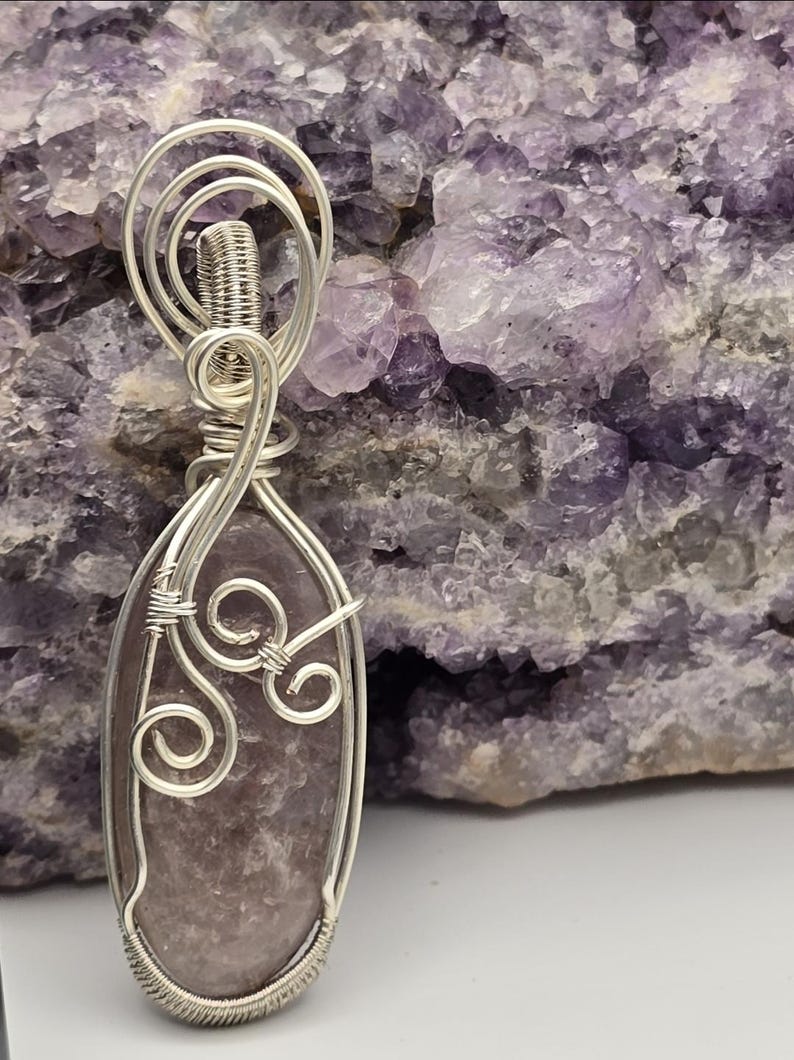 May include: A handcrafted pendant featuring a polished, oval-shaped, lavender-colored stone encased in a silver wire frame. The wirework includes decorative swirls and a wrapped top, creating a unique jewelry piece. The background is a purple crystal formation.