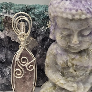May include: A handcrafted pendant with a lavender-colored stone wrapped in silver wire. The pendant is displayed next to a carved purple Buddha statue. The background features a rough, textured green and purple stone.