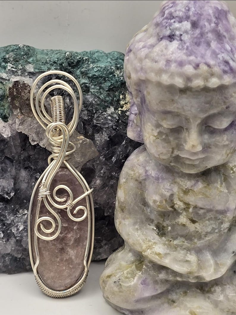 May include: A handcrafted pendant with a lavender-colored stone wrapped in silver wire. The pendant is displayed next to a carved purple Buddha statue. The background features a rough, textured green and purple stone.