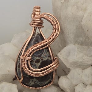 May include: A handcrafted pendant featuring a teardrop-shaped stone with a mottled pattern of dark brown and cream colors. The stone is encased in copper wire, intricately woven to create a decorative frame and bail for hanging.