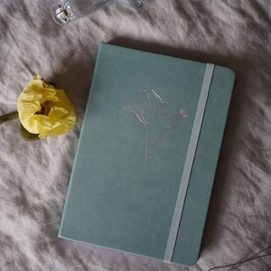 Könnte beinhalten: Ein blaugrünes Notizbuch mit einem silbernen Mohnblumen-Design und einem weißen Gummibandverschluss. Das Notizbuch liegt auf einem weißen Leinenstoff-Hintergrund.
