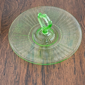 May include: A vintage, light green glass serving tray with a central handle. The tray features a circular design with etched floral patterns and radial lines. The handle is arched and detailed. The tray is approximately 10 inches in diameter.
