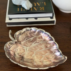 May include: A silver-toned leaf-shaped dish with intricate veining details. The dish has a pearlescent sheen, reflecting shades of pink and purple. The decorative piece is likely used for holding small items or as a display accent.