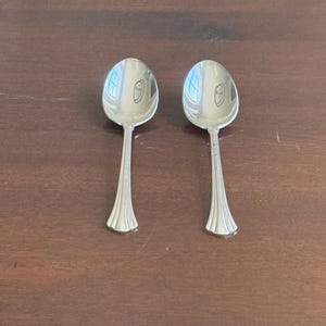 May include: Two polished silver-toned metal spoons with oval bowls and handles that feature a flared base with vertical grooved detailing. The spoons have a reflective surface showing light reflections and a simple, clean design for dining use.