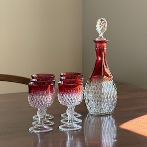 May include: A glass decanter with a faceted stopper and four matching stemmed glasses. Each piece features a clear diamond-cut pattern on the lower half and a deep red glass finish on the upper rim and neck, creating a coordinated set of glassware.